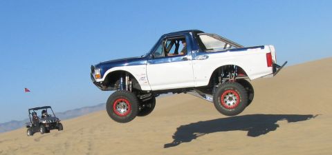 1984 Ford Bronco - A heavily modified Ford Bronco jumps over sand dunes, with a smaller vehicle visible in the distance.