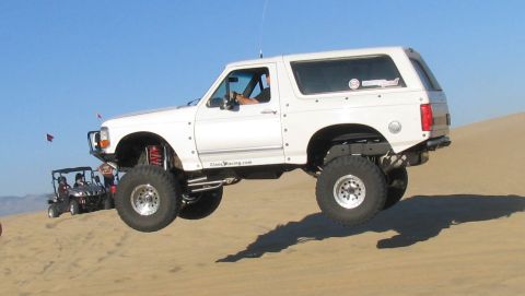1984 Ford Bronco - A white Ford Bronco is captured mid-air as it jumps over sand dunes, with a golf cart visible in the background.