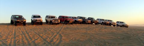 1984 Ford Bronco - A convoy of off-road vehicles is lined up on a sandy slope at sunset.