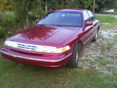 1995 Ford Crown Victoria - A red 1995 Ford Crown Victoria is parked on a grassy area with trees behind it.