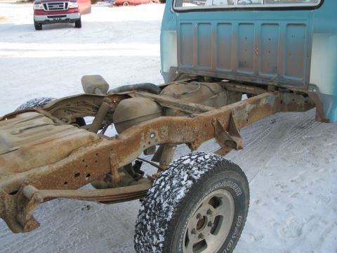 1982 Ford F-150 - The rear chassis of a blue truck is visible, showing significant rust on the frame and a snow-covered tire. Another red truck is in the bac…