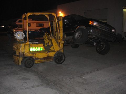 1989 Ford Bronco - A forklift is lifting a black pickup truck, with an RV visible in the background at night.