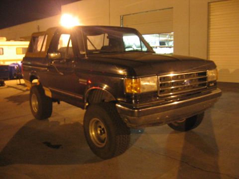 1989 Ford Bronco - A black Ford Bronco with a tan soft top is parked in front of a garage door at night.