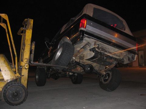 1989 Ford Bronco - Forklift lifting a 1989 Ford Bronco by its frame, showing the rear underside.