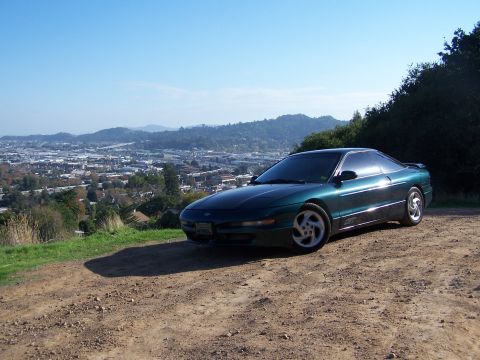 1996 Ford Probe - A green coupe is parked on a dirt road with a city in the background.