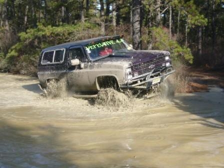 1987 Suzuki Samurai - Off-road vehicle navigates a muddy trail, kicking up water and dirt.