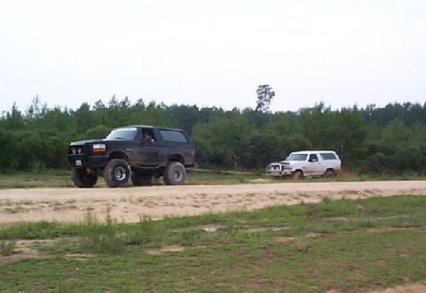 1995 Ford Bronco - Two Ford Broncos are seen on a dirt road, with one towing the other through a wooded area.