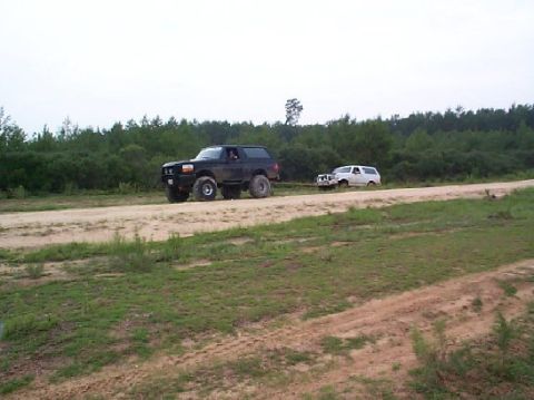 1995 Ford Bronco - A black 1995 Ford Bronco tows a white SUV on a dirt path through a wooded area.