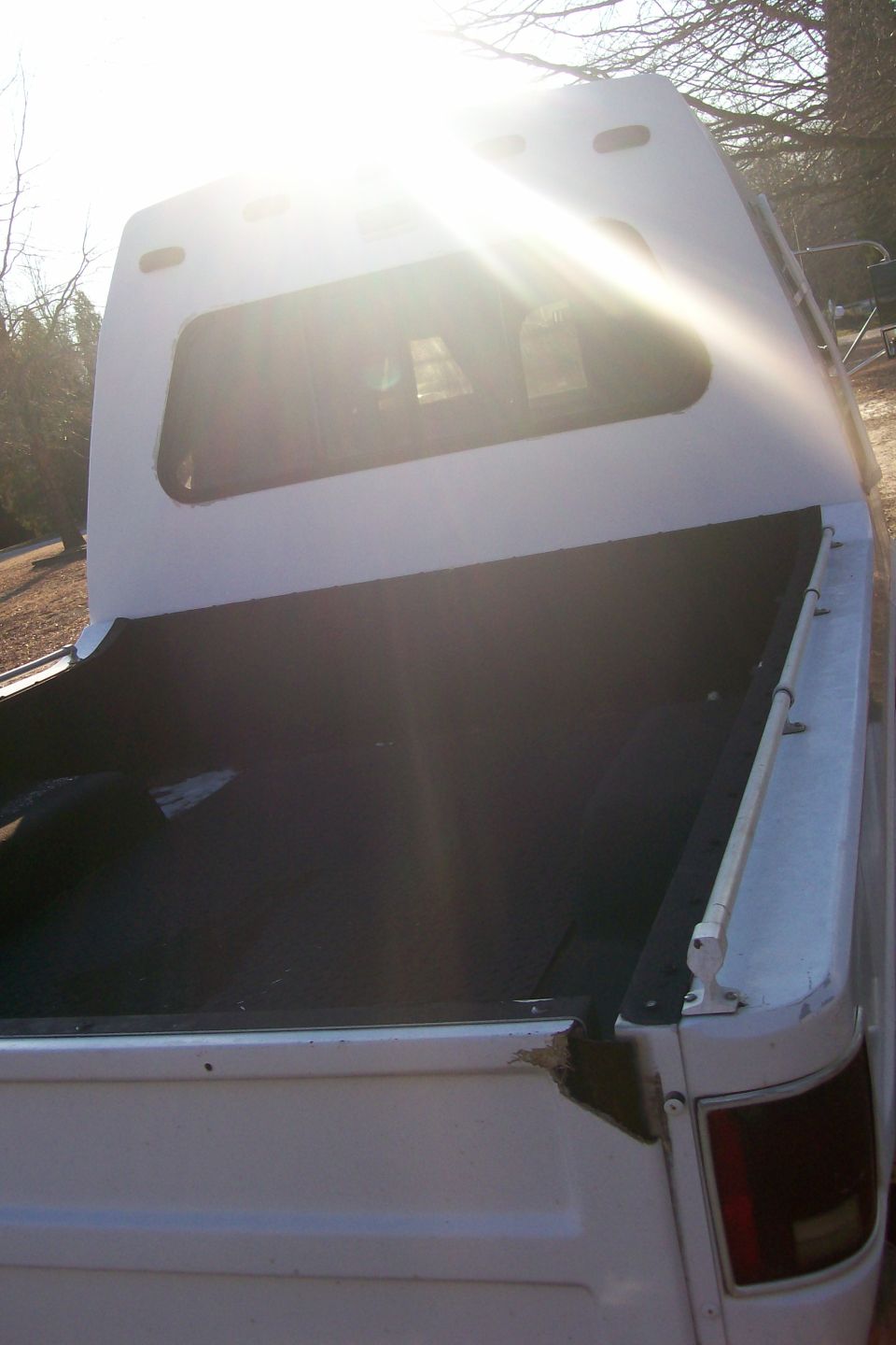 Rear view of a white pickup truck with a camper shell, sun glare, black bed liner, and white side rail.