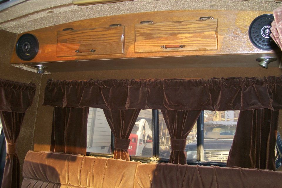 Interior view of a van with wood cabinets, speakers, and brown velvet curtains. Another vehicle is visible through the window