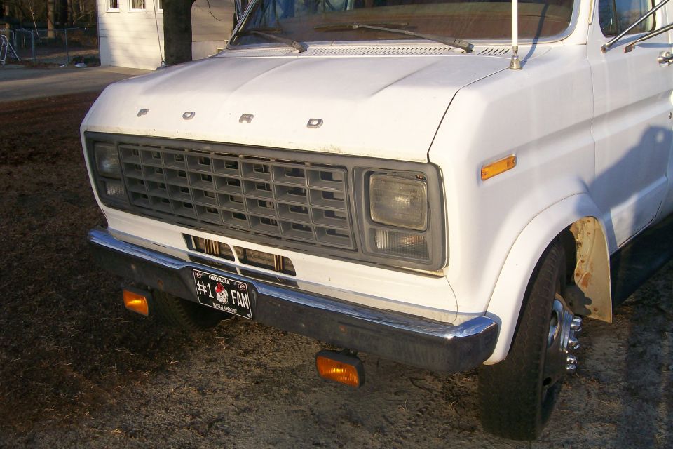 Front view of a white Ford Econoline van. It has a grey grille and a "#1 FAN" license plate