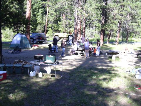 1991 Ford Bronco - A dark colored SUV is parked at a campsite next to a tent and a yellow off-road vehicle. Several people are gathered around picnic tables a…