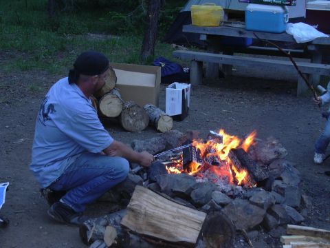 1991 Ford Bronco - A man is tending a campfire in a campsite setting. He is wearing a blue striped shirt and jeans.