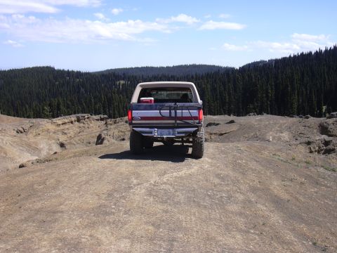 1991 Ford Bronco - The rear view of a lifted, older model Ford Bronco is shown parked on a dirt road with a dense forest in the background. The vehicle has a…