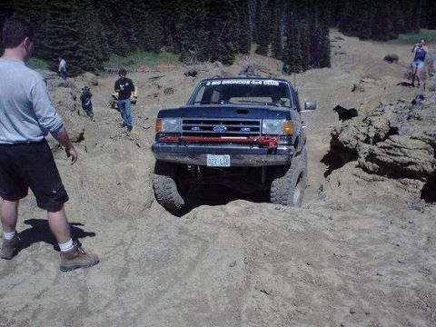 1991 Ford Bronco - A dark blue Ford Bronco is navigating a rocky, off-road trail. Several people are visible in the background, observing the vehicle.