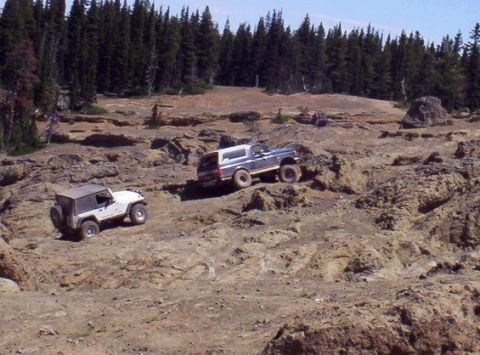 1991 Ford Bronco - Two off-road vehicles navigate a rocky, uneven terrain with a dense forest in the background. A white Jeep is positioned lower on the slope…