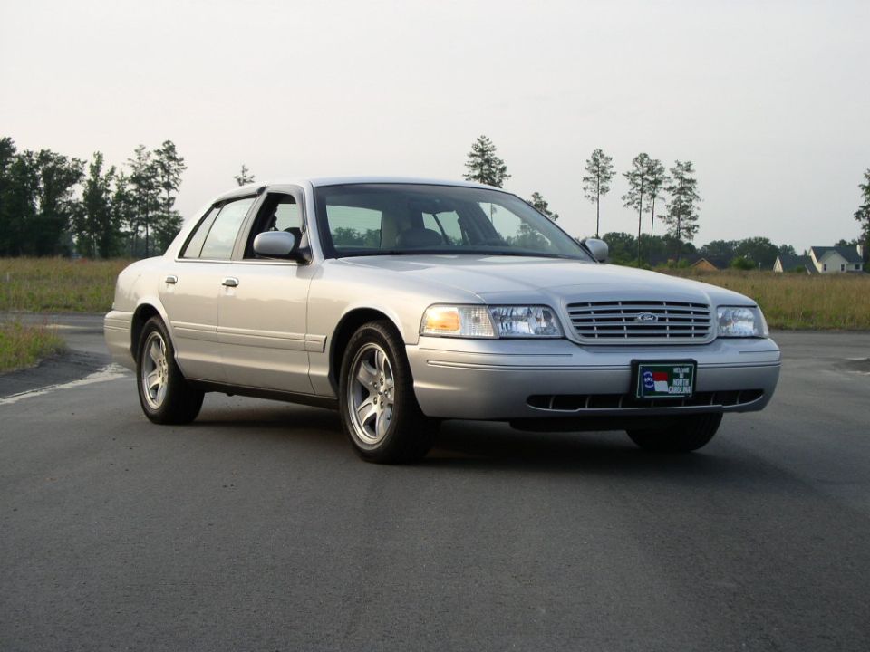 Silver 2001 Ford Crown Victoria sedan parked on asphalt road, trees and houses in background