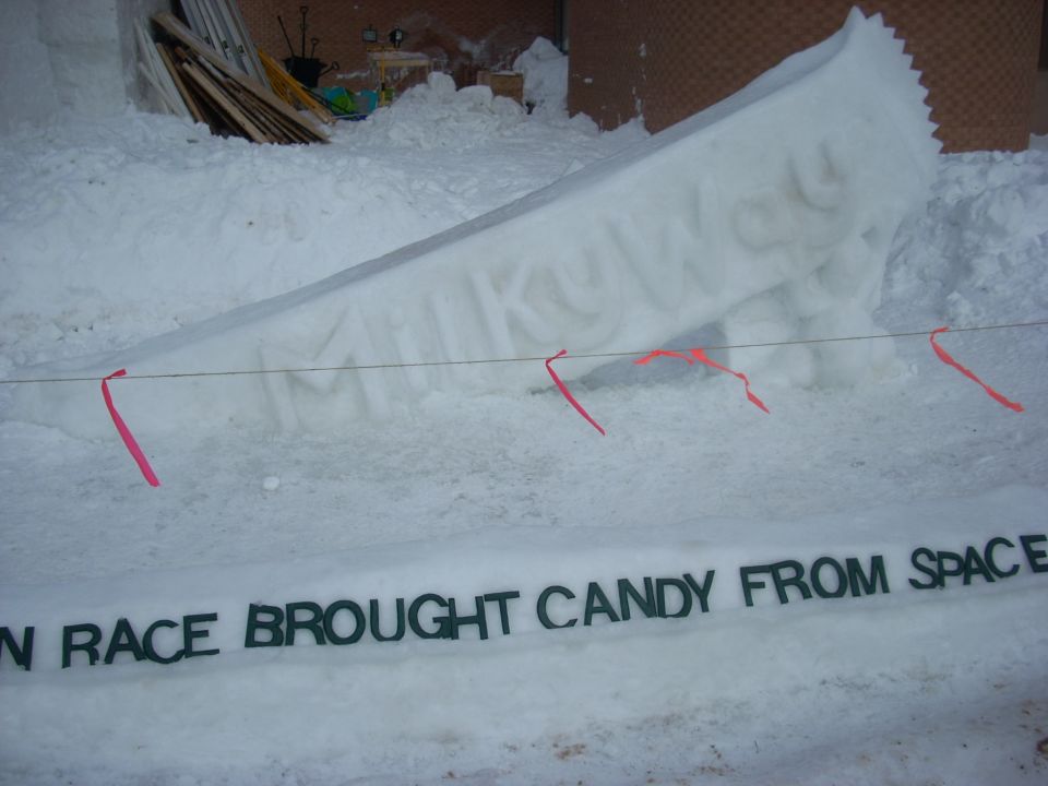 Snow sculpture of a Milky Way candy bar with text "N RACE BROUGHT CANDY FROM SPACE" in front.