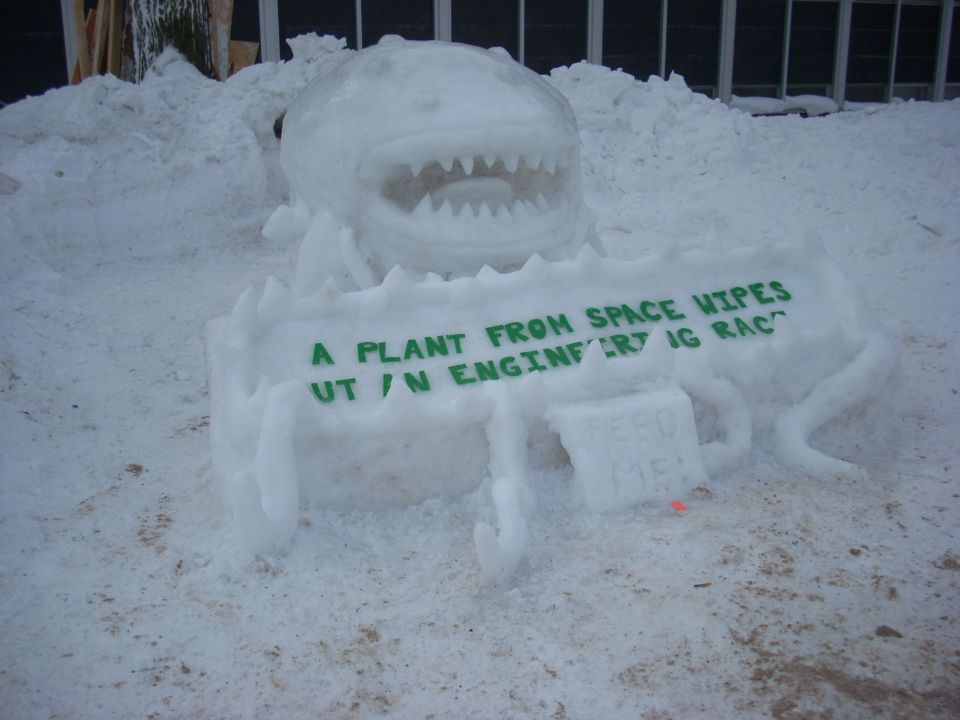 Snow sculpture of a creature with sharp teeth and a sign that reads "A PLANT FROM SPACE WIPES UT / N ENGINFERT IG RAC" and "FEED ME!"