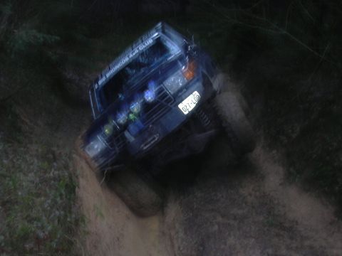 1991 Ford Bronco - A dark blue 1991 Ford Bronco is shown driving off-road on a dirt path at night. The vehicle has off-road lights mounted on the roof and a l…