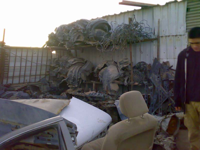Scrap metal and car parts piled outdoors with a person standing to the right.
