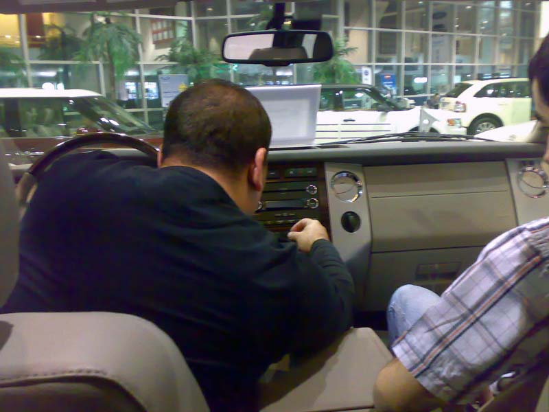Two guys inside a car, one looking at a laptop on the dash. Other cars visible through the windows
