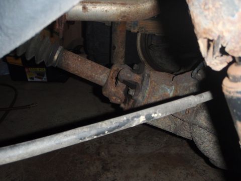 1996 Ford Bronco - The underside of a vehicle shows a rusty driveshaft with a universal joint and a protective rubber boot. A metal bar runs horizontally acro…