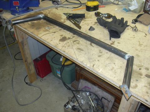 1991 Ford Bronco - Shop-fabricated tubular steel hoop or frame section laid out on a plywood workbench, with welded joints and flat mounting tabs at the corne…