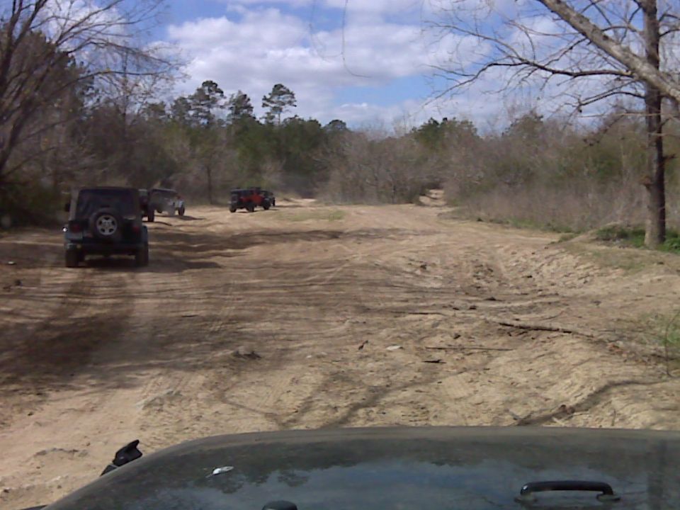 Black Jeep Wrangler with spare tire on rear, followed by other Jeeps on a dirt road