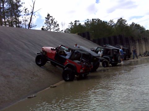 2008 Jeep Wrangler - A red 2008 Jeep Wrangler navigates a steep, wet embankment, followed by other off-road vehicles.
