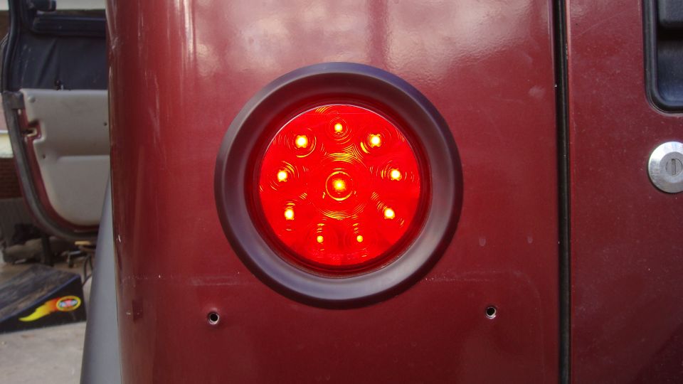 Close-up of a round red LED tail light on a dark red vehicle. Part of the door and interior are visible on the left