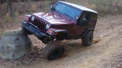 1995 Ford Bronco - A maroon Jeep Wrangler TJ Sahara with a black hardtop flexes off a dirt bank beside a concrete culvert section, right front tire drooped. I…