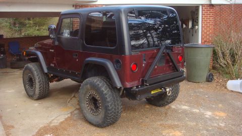 1995 Ford Bronco - Three-quarter rear view of a lifted maroon Jeep Wrangler with a black hardtop, flat fender flares and a black tube rear bumper with tire ca…