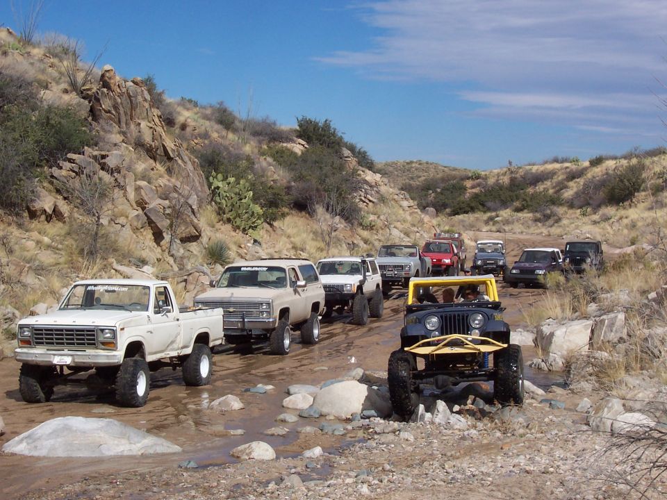Off-road convoy led by a white Ford F-150 pickup truck driving through a rocky creek bed.