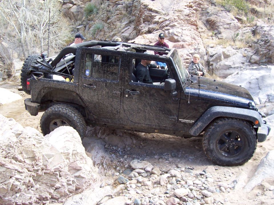 Mud-covered black Jeep Wrangler driving through a rocky, dry riverbed with people.
