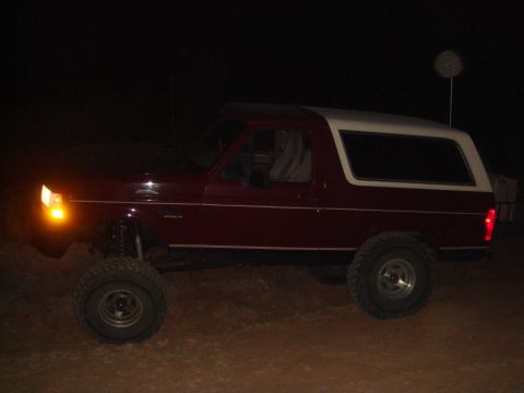 1992 Ford Bronco - A dark red 1992 Ford Bronco with a white top is parked on dirt at night. The front turn signal is illuminated, and the vehicle has large of…