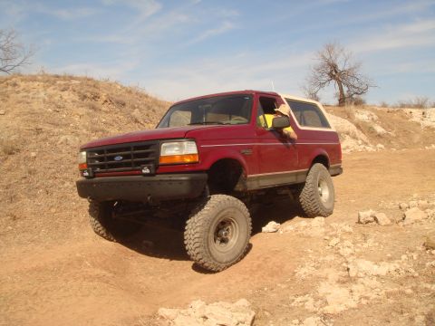 1992 Ford Bronco - A red 1992 Ford Bronco is parked on a dirt road with large off-road tires. The vehicle is angled towards the viewer with the driver's side…