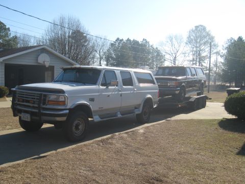 1988 Ford C-350 (Centurion Conversions) - A white Ford extended SUV tows a dark Ford extended SUV on a trailer. The front vehicle's body style suggests a 1992 model year.