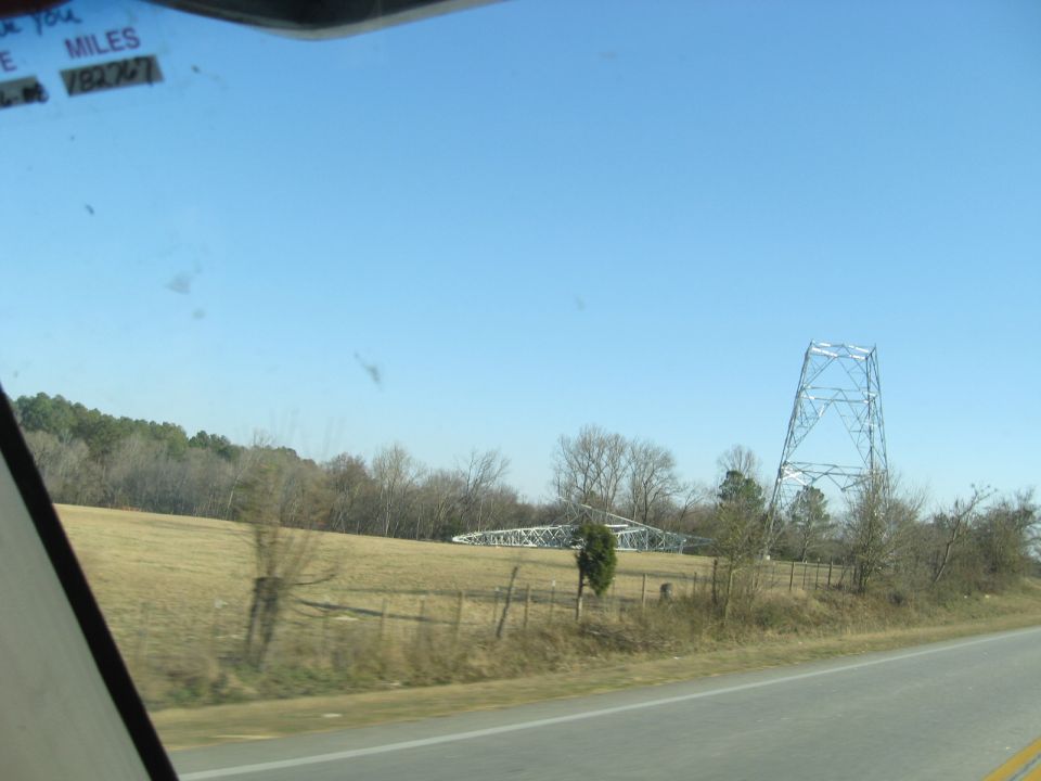 Fallen metal power line tower in a field next to a road, with a standing tower in the background