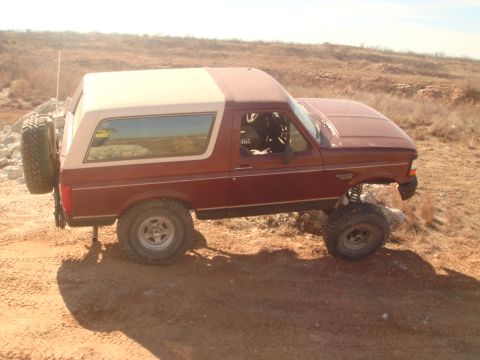 1992 Ford Bronco - The side profile of a maroon and tan Ford Bronco is shown parked on a dirt path in a dry, hilly landscape. The vehicle has large off-road t…