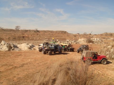 1992 Ford Bronco - A group of off-road vehicles are parked in a dirt lot with a hilly, arid landscape in the background. A red Jeep Wrangler is in the foregro…