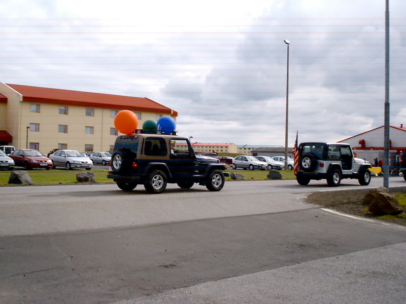 Dark SUV with roof balloons and rear spare tire on road, white Jeep behind