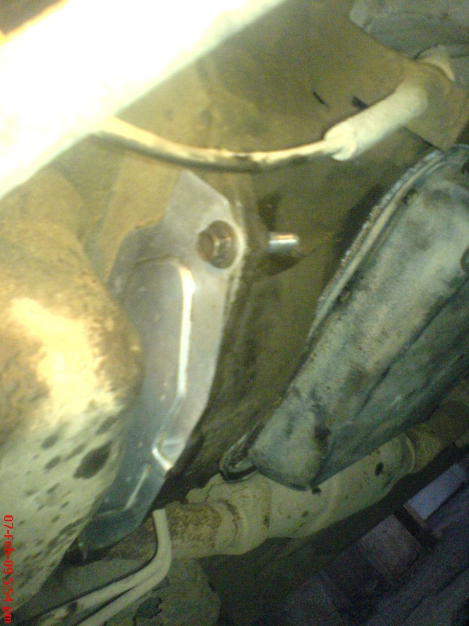 Underside view of car showing oil pan drain plug, oily pan surface and exhaust piping