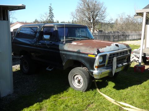 1991 Ford Bronco - A black Ford Bronco with a rusty hood is parked on a grassy lawn next to a white structure. The vehicle is viewed from a 3/4 front angle, s…