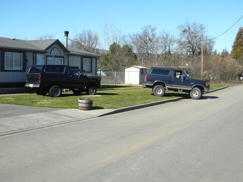 1991 Ford Bronco - Two dark-colored Ford Broncos are parked on a grassy area next to a paved road. The Bronco on the left is parked in front of a house, while…