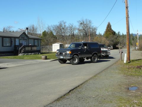 1991 Ford Bronco - A black Ford Bronco is parked on a paved road in front of a house, with another white and red truck behind it. The Bronco has large off-roa…