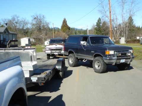 1991 Ford Bronco - A dark blue Ford Bronco is parked on a paved road, facing right, with a white trailer attached. Another older white pickup truck is visible…