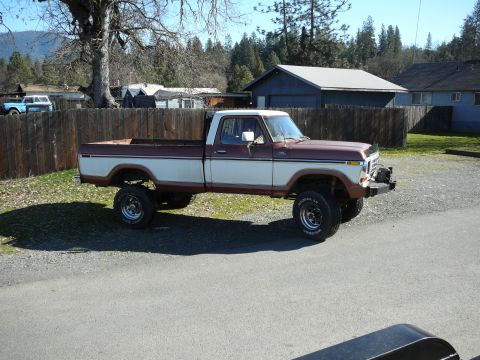 1991 Ford Bronco - A lifted, two-tone brown and white 1970s era pickup truck is parked on a gravel lot next to a wooden fence. The truck has large off-road ti…