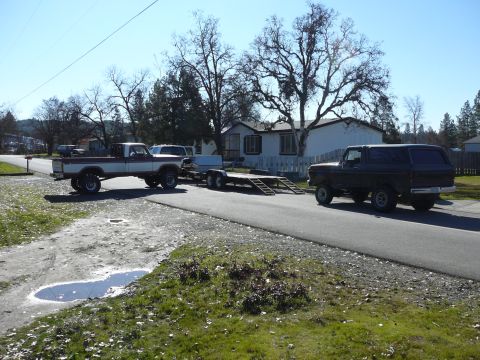 1991 Ford Bronco - A lifted, two-tone pickup truck is parked on a gravel road, towing a trailer. A dark-colored SUV is driving down the adjacent paved road.