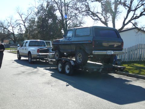1991 Ford Bronco - A dark-colored Ford Bronco is loaded onto a car trailer towed by a white Ford pickup truck. The Bronco is positioned facing forward on the…